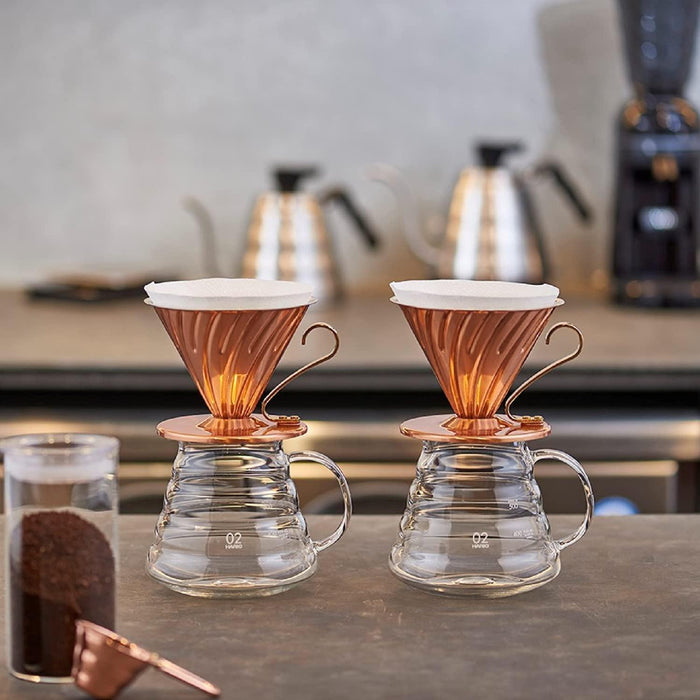 Hario v60 copper drippers side by side on counter top with kettles in the background. Coffee ground in jar next to them